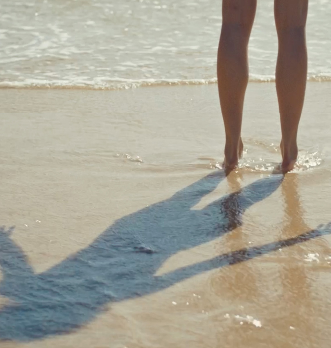 A person walks barefoot through shallow water on a beach.