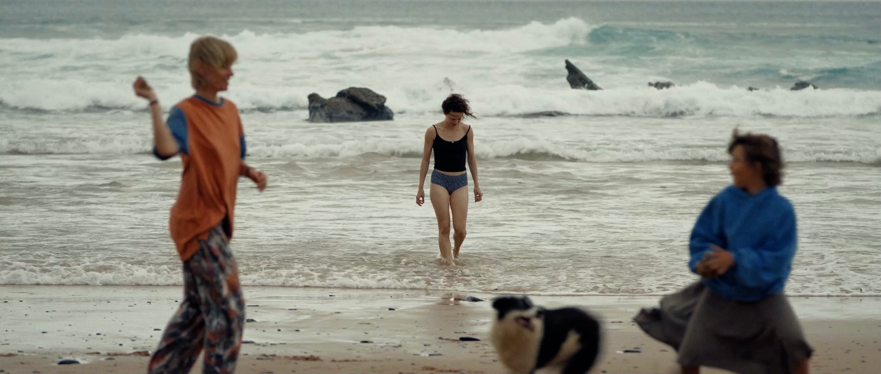 A young woman comes out of the surf as people and a dog cross the beach.